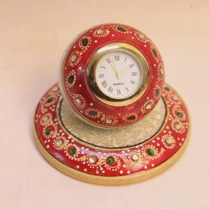 Puede incluir: Un reloj de cuarzo decorativo con una esfera redonda roja y dorada. El reloj está colocado en una base roja y dorada a juego, adornada con piedras decorativas. La esfera del reloj tiene números romanos y agujas doradas.