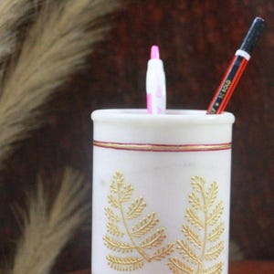 Puede incluir: Porta bolígrafos de mármol blanco con diseño de hojas doradas y ribete rojo y dorado. El portalápices contiene un bolígrafo rosa y un lápiz rojo y negro. El diseño presenta dos patrones de hojas de helecho doradas.