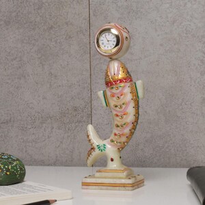 May include: A decorative marble clock in the shape of a fish. The clock has a white face with black Roman numerals and gold hands. The fish is white with gold, pink, and green accents and small decorative jewels. The clock is approximately 10 inches tall.