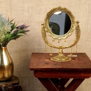 May include: An antique-style, gold-coloured vanity mirror with a circular, scalloped frame. The mirror is supported by a decorative stand and a curved arm with dangling charms. It rests on a small, dark wooden table.