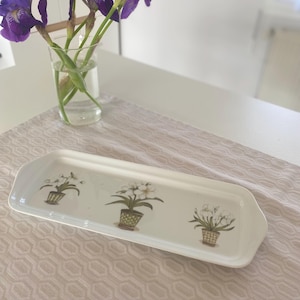 Peut inclure: Plateau de service rectangulaire blanc avec un motif floral. Le plateau présente trois fleurs blanches dans des pots verts avec des accents bruns. Le plateau est sur une surface brun clair.