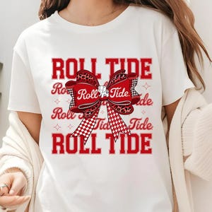 Könnte beinhalten: Weißes T-Shirt mit dem Text "ROLL TIDE" in Rot, mit einem Schleifendesign mit Leopardenmuster und einem rot-weiß karierten Muster. Das Shirt ist ein lässiger Stil.