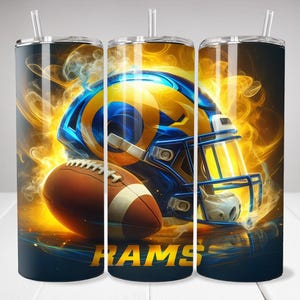 Może przedstawiać: Zestaw trzech kubków ze stali nierdzewnej o tematyce futbolowej. Każdy kubek przedstawia niebiesko-żółty kask futbolowy i brązową piłkę futbolową na ognistym tle. U dołu widnieje napis "RAMS".