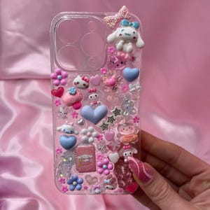 Può includere: Custodia per telefono trasparente decorata con vari ciondoli rosa, blu e bianchi. I ciondoli includono cuori, fiori, stelle e personaggi dei cartoni animati. Sono presenti anche un fiocco rosa e un piccolo taccuino.