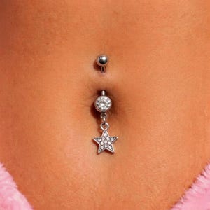 May include: A silver belly button ring featuring a dangling star pendant. The star is adorned with small, clear gemstones. The top of the ring has a small, round silver ball. The jewellery is set against a skin tone background.