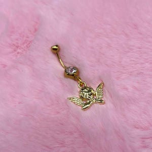 May include: Gold-colored belly button ring featuring a curved barbell design. The top ball and the charm are gold. The charm is a small angel with wings. A clear gem is set in the middle of the barbell.