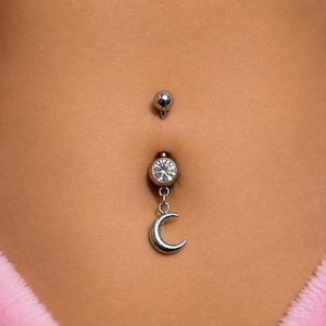 Puede incluir: Una joya de piercing para el ombligo en tono plateado con una bola superior circular y un componente inferior con una piedra preciosa redonda y transparente. Un pequeño dije de media luna plateado y liso cuelga de una argolla metálica.