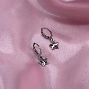 Può includere: Un paio di orecchini a cerchio color argento, ciascuno con un ciondolo a forma di stella pendente. Gli orecchini sono esposti su un tessuto rosa e serico. I ciondoli a stella hanno un design tridimensionale.