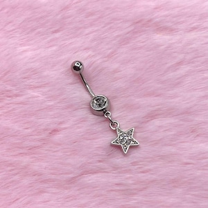 May include: A silver belly button ring with a dangling star charm. The ring features a small, round top ball, a clear gem, and a star-shaped charm encrusted with small, clear gems. The jewelry is set against a pink, fuzzy background.