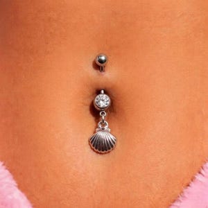 May include: A close-up of a belly button piercing. The silver-coloured piercing has a small, round top ball, a clear gem, and a dangling seashell charm. The skin is a light tan colour.