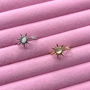 May include: Two rings with sunburst designs. One ring is silver-toned with a white stone center and a small clear stone accent. The other ring is gold-toned with a similar design and a light-colored stone center and a small clear stone accent.
