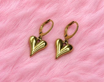 Puede incluir: Un par de pendientes de aro dorados con colgantes tridimensionales en forma de corazón. Los corazones tienen un acabado metálico liso y pulido con una sutil hendidura vertical que recorre el centro de cada una de las piezas.