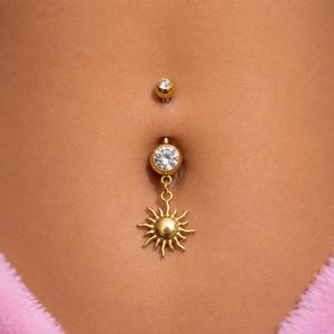 Puede incluir: Un piercing de ombligo en tono dorado que presenta una pequeña piedra transparente en la bola superior y una piedra más grande en la pieza inferior, conectada a un dije colgante con forma de sol y rayos radiantes en acabado dorado.