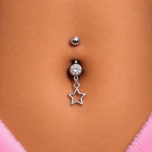 May include: A silver-toned navel ring featuring a circular clear gemstone set in the lower ball, with a dangling open-frame star charm suspended from a small jump ring. The barbell includes a smooth, polished silver-toned top ball.