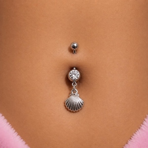 Puede incluir: Un piercing para el ombligo de tono plateado que presenta una pequeña piedra preciosa redonda y transparente facetada en la parte superior del colgante y un dije en forma de concha marina, asegurado por una bola lisa y redonda.