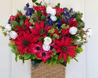 May include: A floral arrangement in a woven basket. The arrangement features red, white, and blue flowers, including large red daisies, white roses, and blue irises, with green foliage. The basket is square and made of a light brown woven material.