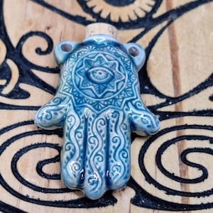 May include: A blue ceramic bottle stopper in the shape of a Hamsa hand, featuring an eye design in the center and intricate patterns. The stopper has a cork base and is set against a wooden background with eye and spiral designs.