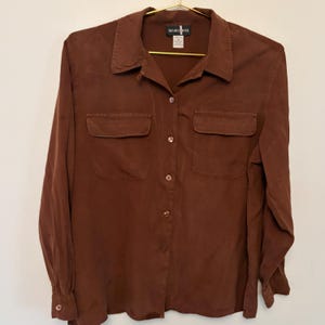 May include: A brown button-down shirt with two front pockets. The long-sleeved shirt has a collared neckline and is hanging on a gold hanger. The tag reads "SILK NICOLETER".