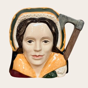 May include: A ceramic character jug shaped as a person with dark hair, wearing a green and yellow bonnet and a yellow collar. The handle of the jug is designed to look like a wooden-handled axe with a grey metal blade, extending from the right side.