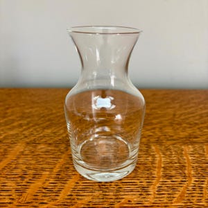 May include: A clear glass vase with a wide base and a flared top. The vase is empty and sits on a wooden surface. The glass is transparent, allowing light to pass through.