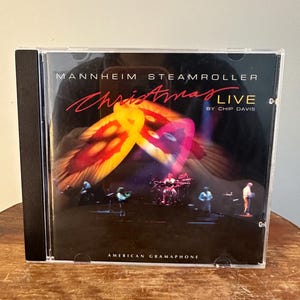 Op de afbeelding: Een cd-hoesje voor het album "Christmas Live" van Mannheim Steamroller door Chip Davis. De cover heeft een levendig, abstract ontwerp in tinten rood, oranje en geel, met een band die optreedt op een donker podium. De tekst "Christmas Live" is rood.