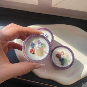 Könnte beinhalten: Drei kleine violette Kerzenhalter mit weißen Kerzen und bunten Steinen, die in das Wachs eingebettet sind. Die Kerzen stehen auf einer weißen Oberfläche.