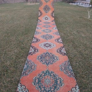May include: An orange runner rug with a repeating geometric pattern in teal, navy, and cream. The rug is displayed outdoors on a grassy lawn, showcasing its length and intricate design. The rug's border features a detailed, dark pattern.