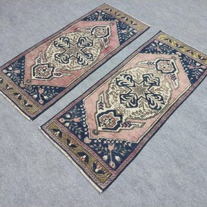 May include: Two rectangular rugs with detailed patterns. The rugs have a central design in cream and pink, framed by a navy blue border with floral motifs. The edges are finished with a gold and green trim. The rugs are placed side by side.