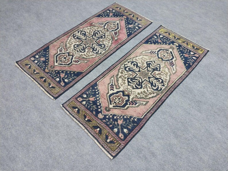 May include: Two rectangular rugs with detailed patterns. The rugs have a central design in cream and pink, framed by a navy blue border with floral motifs. The edges are finished with a gold and green trim. The rugs are placed side by side.