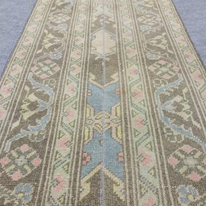 May include: A long, rectangular rug with a detailed, symmetrical pattern. The rug features a central design in shades of brown, beige, and blue, with floral and geometric motifs. Pink and light green accents add pops of colour to the design.