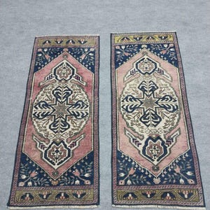 May include: Two rectangular rugs with detailed patterns. Each rug has a central cream and navy blue design, framed by a pink border and navy blue corners. The rugs display a traditional, ornate style.