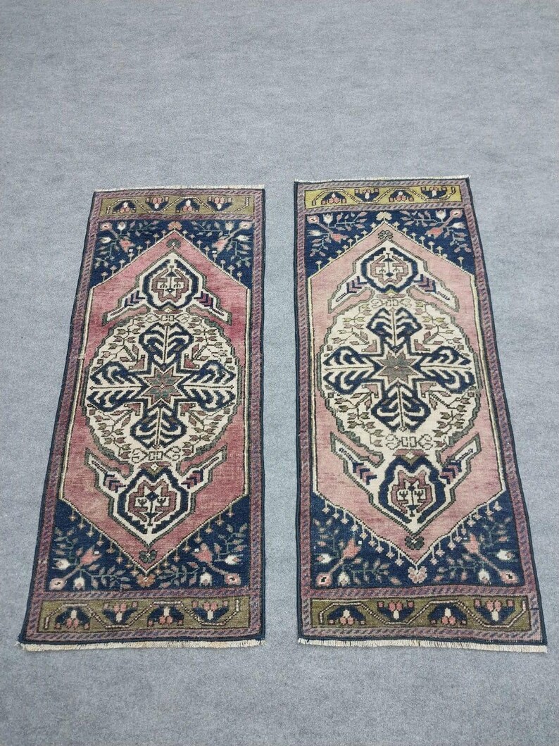 May include: Two rectangular rugs with detailed patterns. Each rug has a central cream and navy blue design, framed by a pink border and navy blue corners. The rugs display a traditional, ornate style.