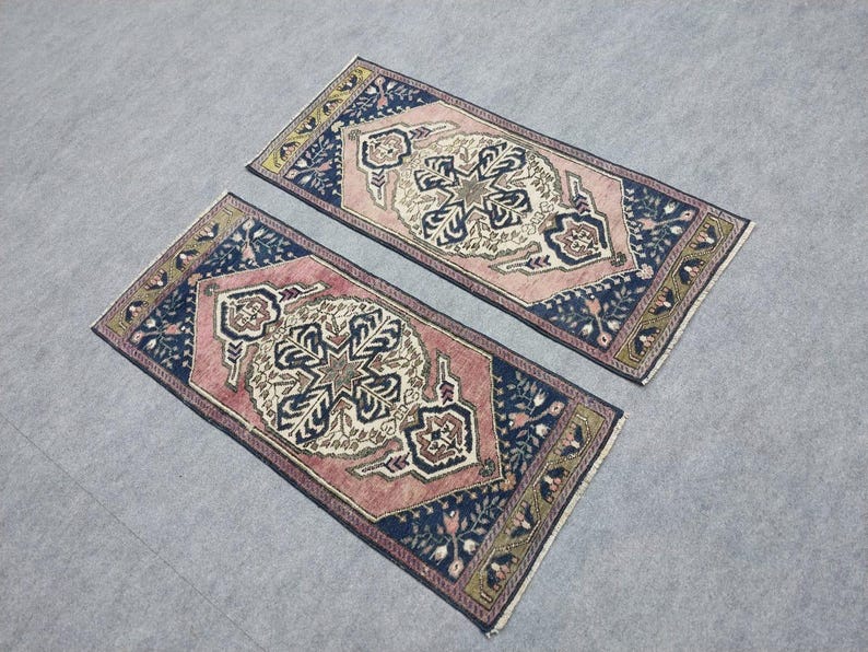 May include: Two rectangular rugs with detailed designs. The rugs have a central cream and brown pattern, framed by a pink border and navy blue corners. The edges are decorated with floral and geometric patterns.