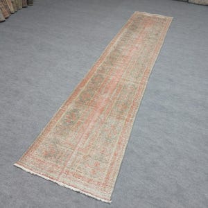 May include: A long, rectangular runner rug with a faded, distressed appearance. The rug features a muted color palette of peach, green, and cream, with a geometric pattern. The rug is likely made of woven material and is suitable for a hallway or entryway.