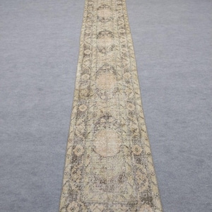 May include: A long, rectangular rug with a detailed, faded design. The rug features a central pattern of repeating medallions in beige, brown, and cream. The edges and background have a distressed appearance, adding to its vintage aesthetic.