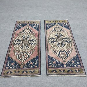 May include: Two rectangular rugs with a central medallion design. The rugs feature a pink field, navy blue borders, and intricate patterns in cream, gold, and navy. The rugs are placed on a grey surface.