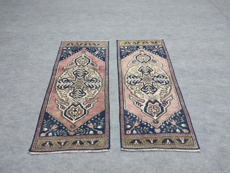 May include: Two rectangular rugs with a central medallion design. The rugs feature a pink field, navy blue borders, and intricate patterns in cream, gold, and navy. The rugs are placed on a grey surface.