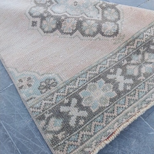 May include: A rectangular woven rug featuring a light beige base with a decorative border and central medallion in muted shades of sage green, soft blue, and charcoal gray. The rug has a textured pile surface and a short fringed edge at the end.