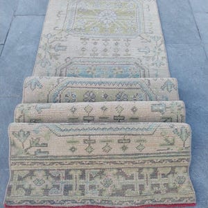 Könnte beinhalten: Ein aufgerollter, rechteckiger Vintage-Teppich mit einem beige-grünen geometrischen Muster. Der Teppich hat einen roten Rand und wird auf einer grauen Oberfläche präsentiert. Das Design umfasst florale und geometrische Elemente.