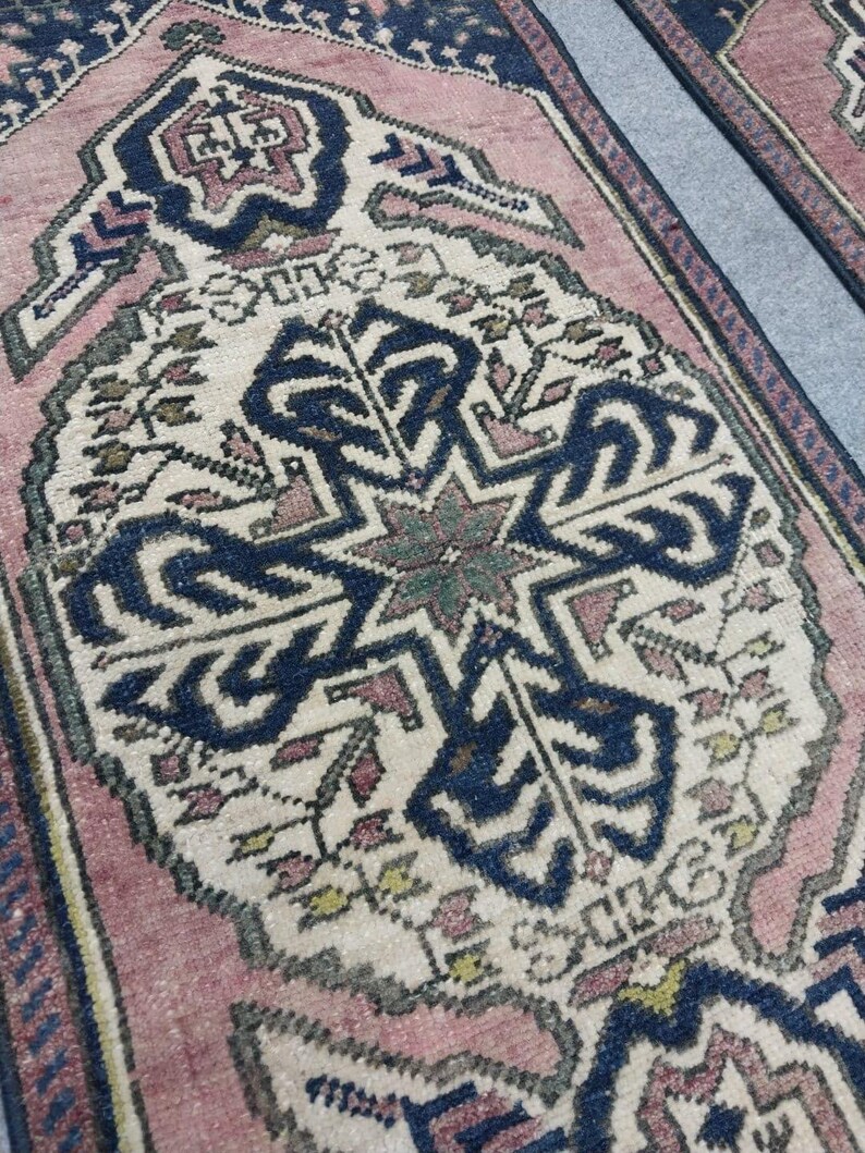 May include: Close-up of two vintage rugs with detailed geometric patterns. The rugs have a central design in navy blue and cream, framed by a pink border. The intricate patterns include floral and star-like motifs, creating a traditional aesthetic.