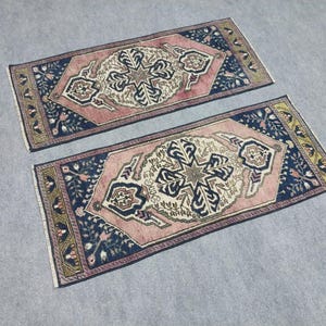 May include: Two rectangular rugs with detailed floral and geometric patterns. The rugs have a central design in cream and navy blue, set against a pink field and a navy blue border. The borders are decorated with floral motifs and a gold trim.