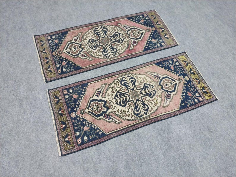 May include: Two rectangular rugs with detailed floral and geometric patterns. The rugs have a central design in cream and navy blue, set against a pink field and a navy blue border. The borders are decorated with floral motifs and a gold trim.