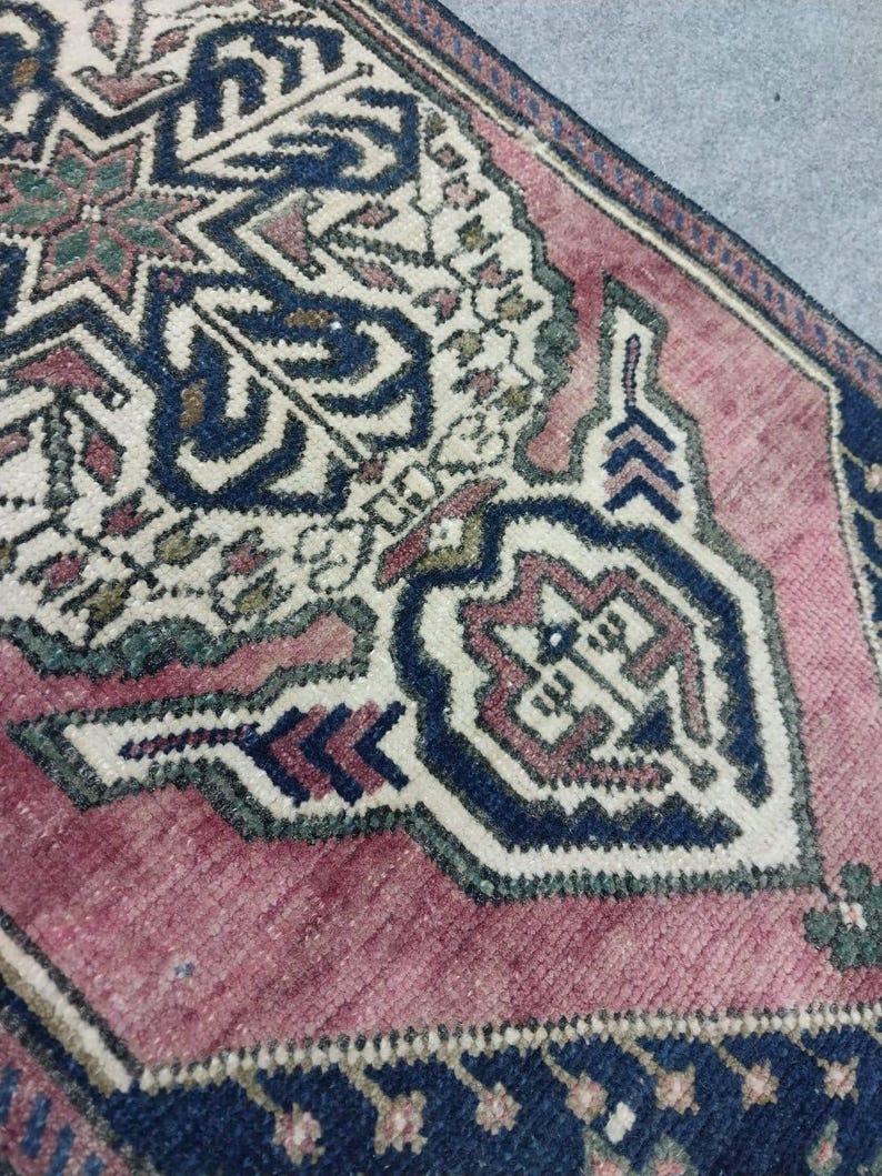 May include: Close-up of a section of a vintage rug with a detailed geometric pattern. The rug features a central design in cream, navy blue, and muted red hues. The border is navy blue with small floral motifs. The rug's texture is visible.