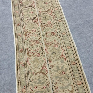 Pode incluir: Um tapete longo e retangular com um intrincado padrão floral. O tapete apresenta um fundo creme com um desenho repetido de flores e folhas em tons de vermelho, castanho e verde. A borda tem um padrão geométrico detalhado.