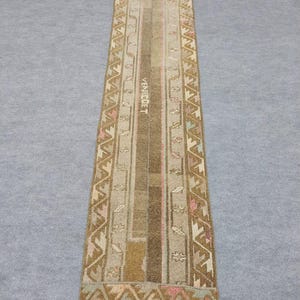 Könnte beinhalten: Ein rechteckiger, vintage Läufer-Teppich mit einem zentralen vertikalen Streifendesign in Braun-, Beige- und Rosatönen. Der Teppich hat geometrische Muster an den Rändern und das Wort "VENECDET" ist sichtbar. Der Teppich ist wahrscheinlich aus gewebtem Material.