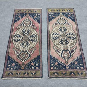 May include: Two rectangular rugs with detailed patterns. Each rug has a central design in cream, pink, and navy blue, framed by a navy border with floral details. The rugs have a traditional, vintage style.