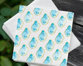 May include: A stack of white paper napkins with a repeating pattern of blue and gold watercolor designs. The design resembles a geode or abstract shape, with a light blue center and gold outline. The napkins are on a dark wooden surface.