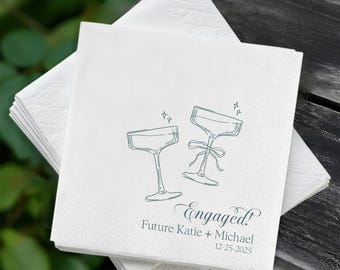 May include: White paper napkins with a teal illustration of two champagne glasses and the word "Engaged!" with the names "Future Katie + Michael" and the date "12-25-2025". The napkins are stacked on top of each other.