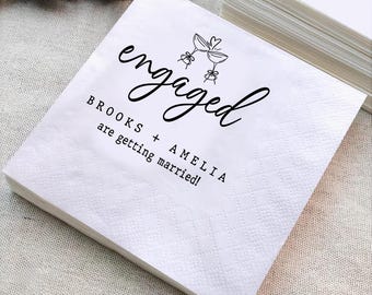 May include: A stack of white paper napkins with a diamond pattern. The word "engaged" is written in cursive script, with a drawing of two champagne glasses. The text "BROOKS + AMELIA are getting married!" is also printed on the napkins.