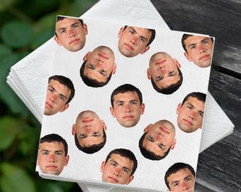 May include: White paper napkins with a repeating pattern of a person's face. The napkins are stacked on top of each other, with the top napkin featuring the face pattern. The background is a blurred green and black.